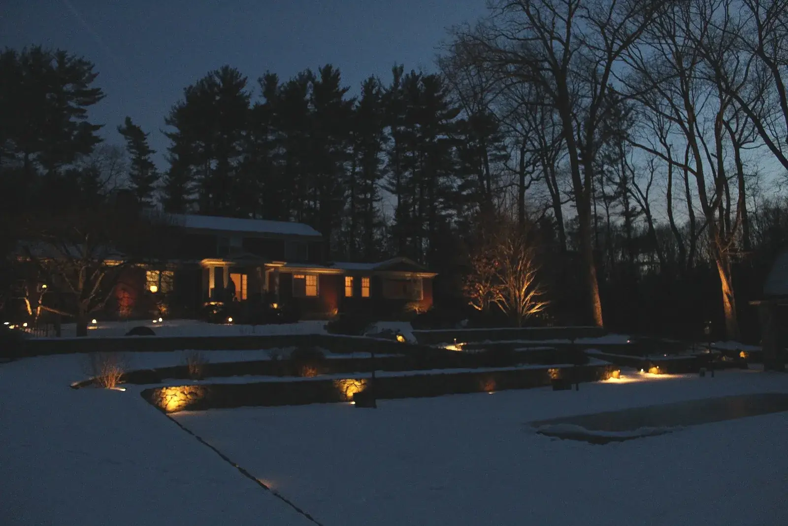 Landscape lighting on a stone wall, walkway, and house at night, with snow on the ground